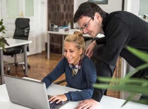 a woman in a blue sweater smiling at a laptop, while a man in glasses looks over her shoulder